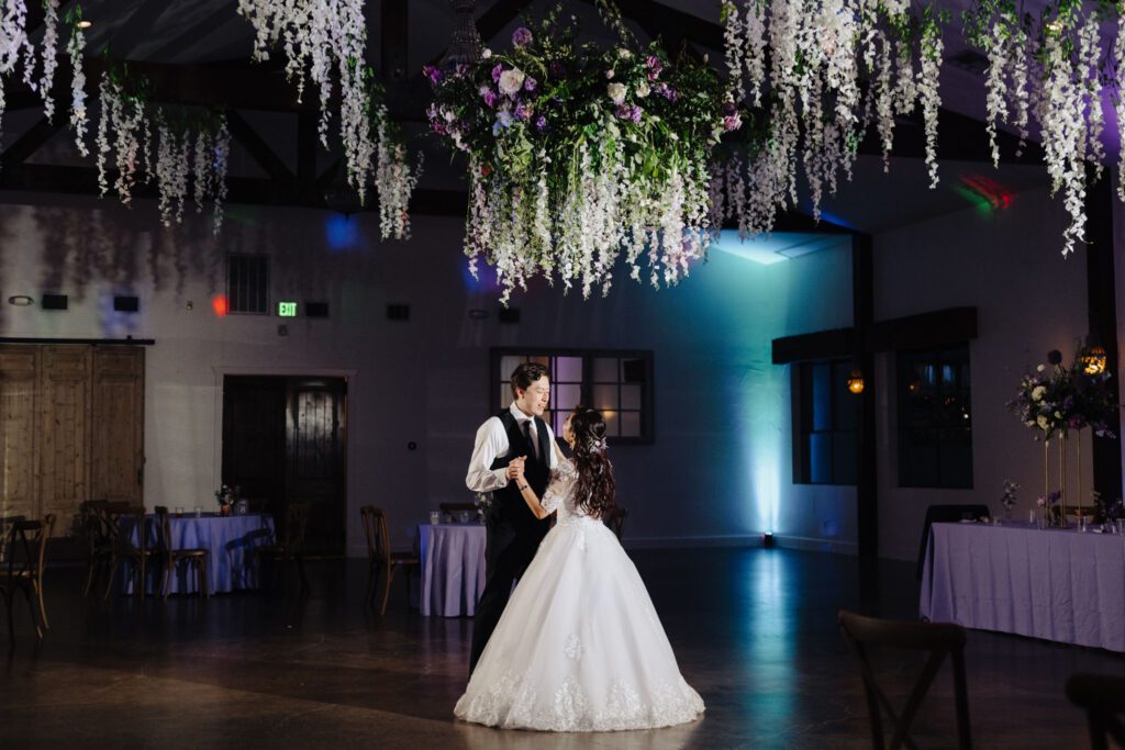 Couple sharing their first dance indoors beneath hanging floral installations and soft lighting