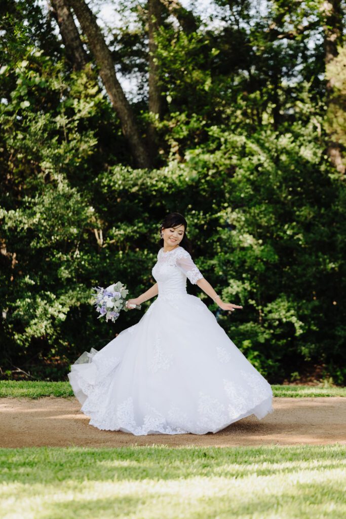 Bride in a white gown twirling outdoors on a tree-lined path holding a bouquet