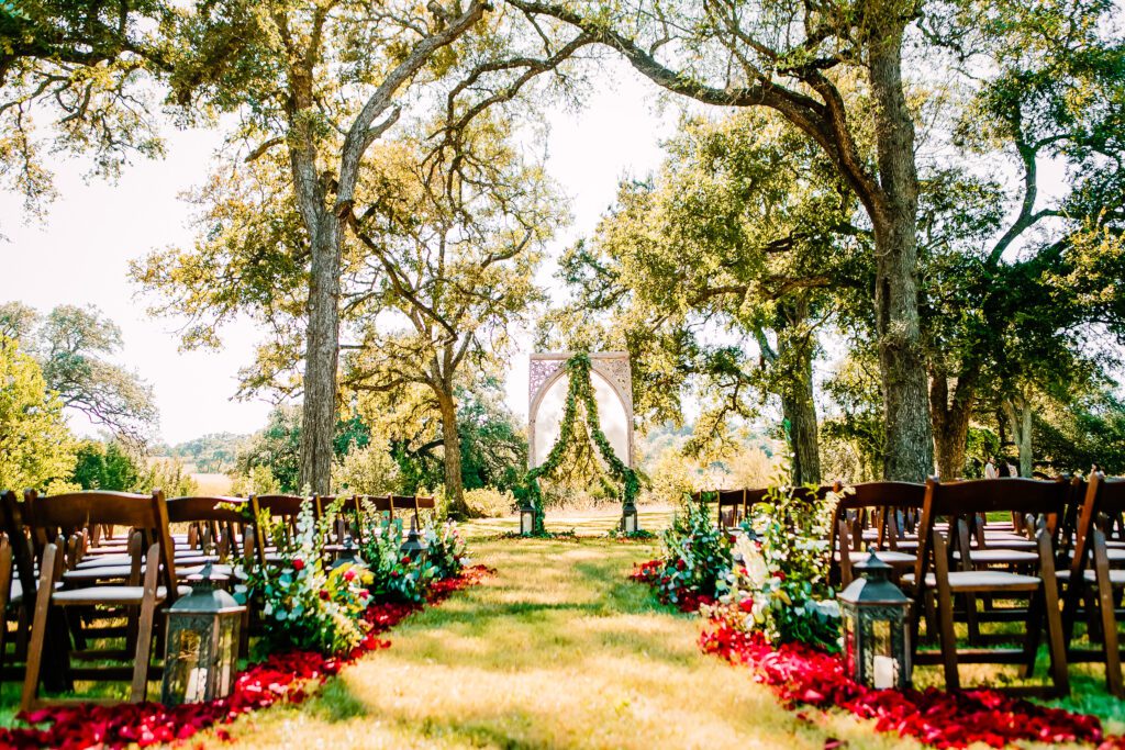Outdoor wedding ceremony aisle lined with red petals and greenery under tall trees