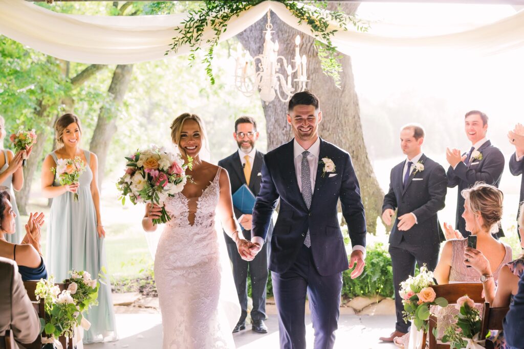 Bride and groom walking down the aisle after ceremony under draped fabric and chandelier with guests applauding