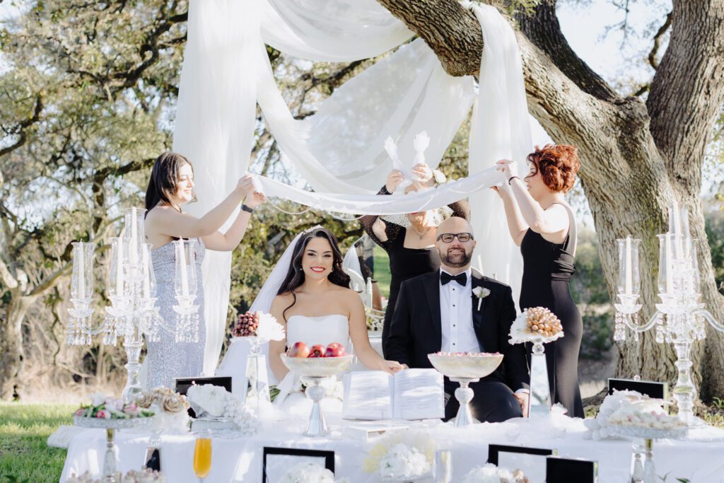 Bride and groom sitting down in front of cakes, and three people fixing cloths above them