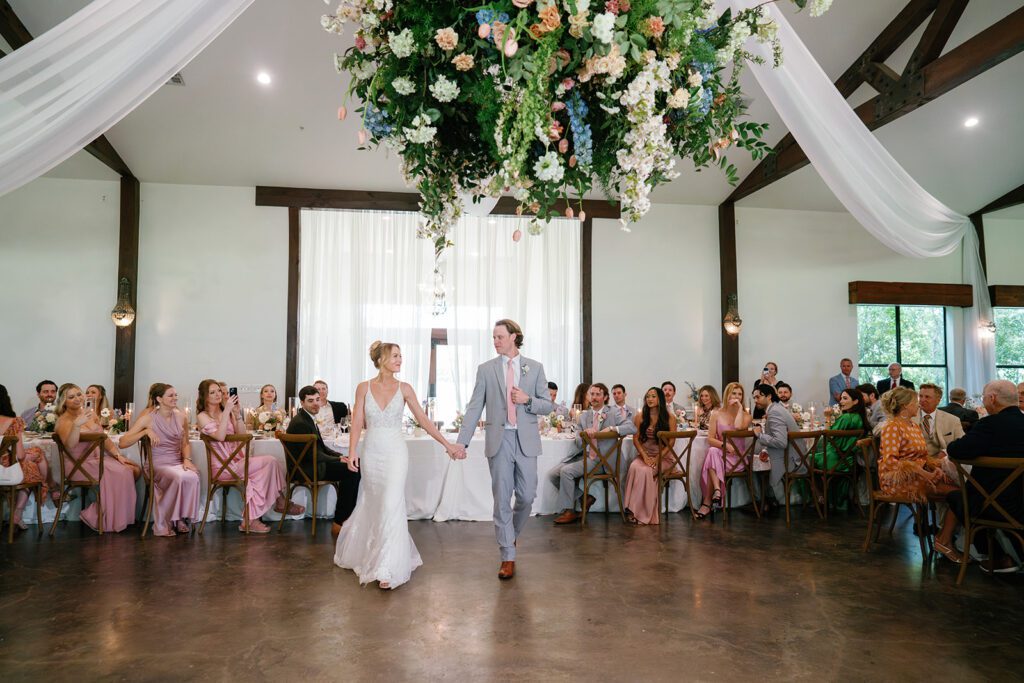 Bride and groom walk hand in hand into their reception under hanging floral arrangements as guests in pastel attire look on