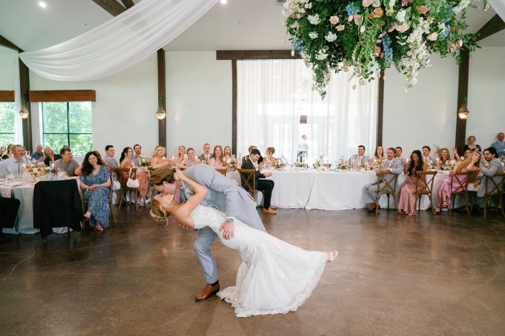 Groom dips the bride during their first dance under a suspended floral installation while guests cheer from their tables
