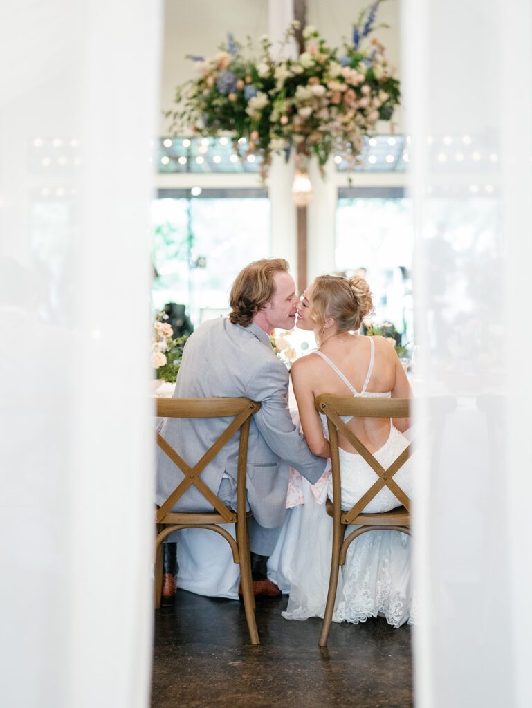 Bride and groom kiss at their reception table, framed by sheer white drapery with a floral installation glowing in the background