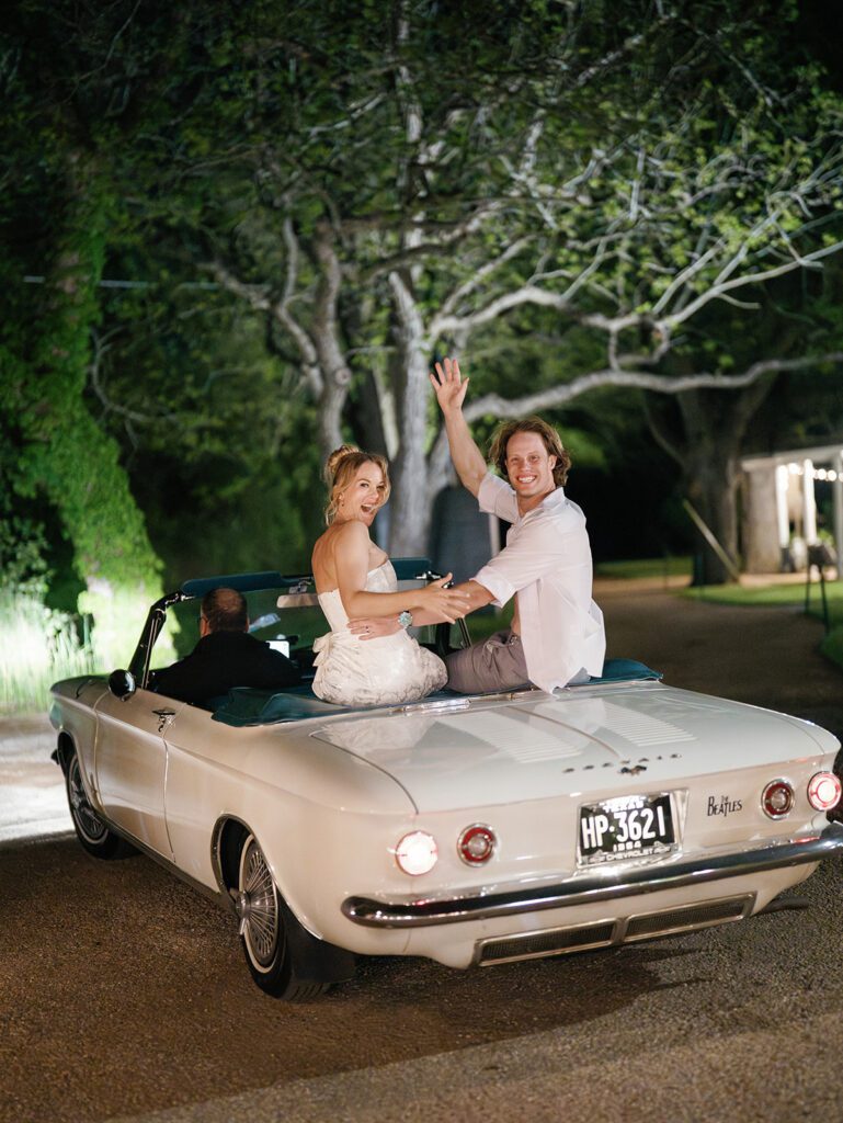 Newlyweds wave from the back of a vintage white Chevrolet Corvair convertible with Texas plates during a nighttime exit under oak trees
