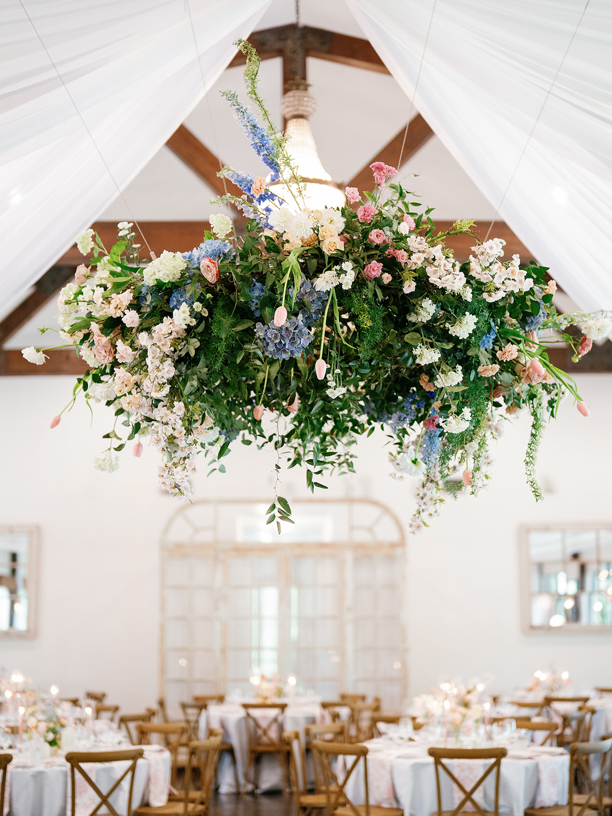 Floral chandelier wedding installation of blue hydrangeas, delphinium, garden roses and pink tulips hanging beneath sheer white ceiling draping