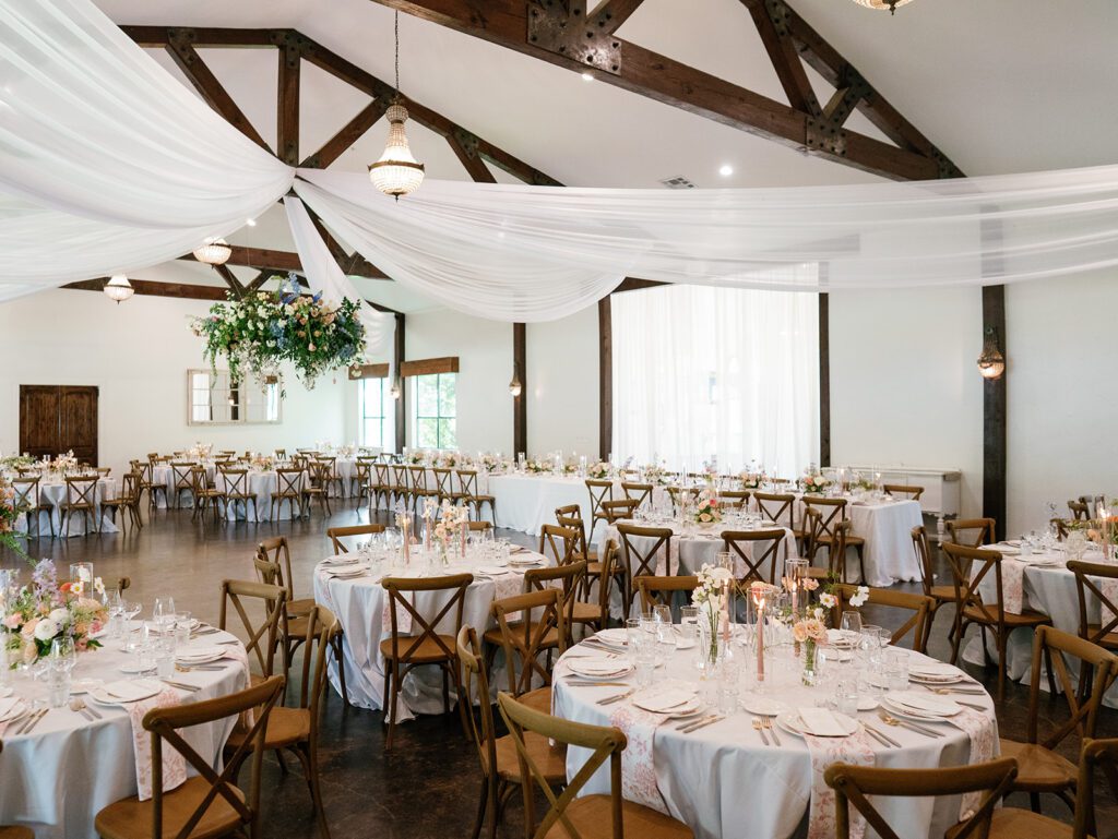 Wide view of a Texas wedding reception hall with exposed wood trusses, white fabric draping, crystal chandeliers and round tables with cross-back chairs