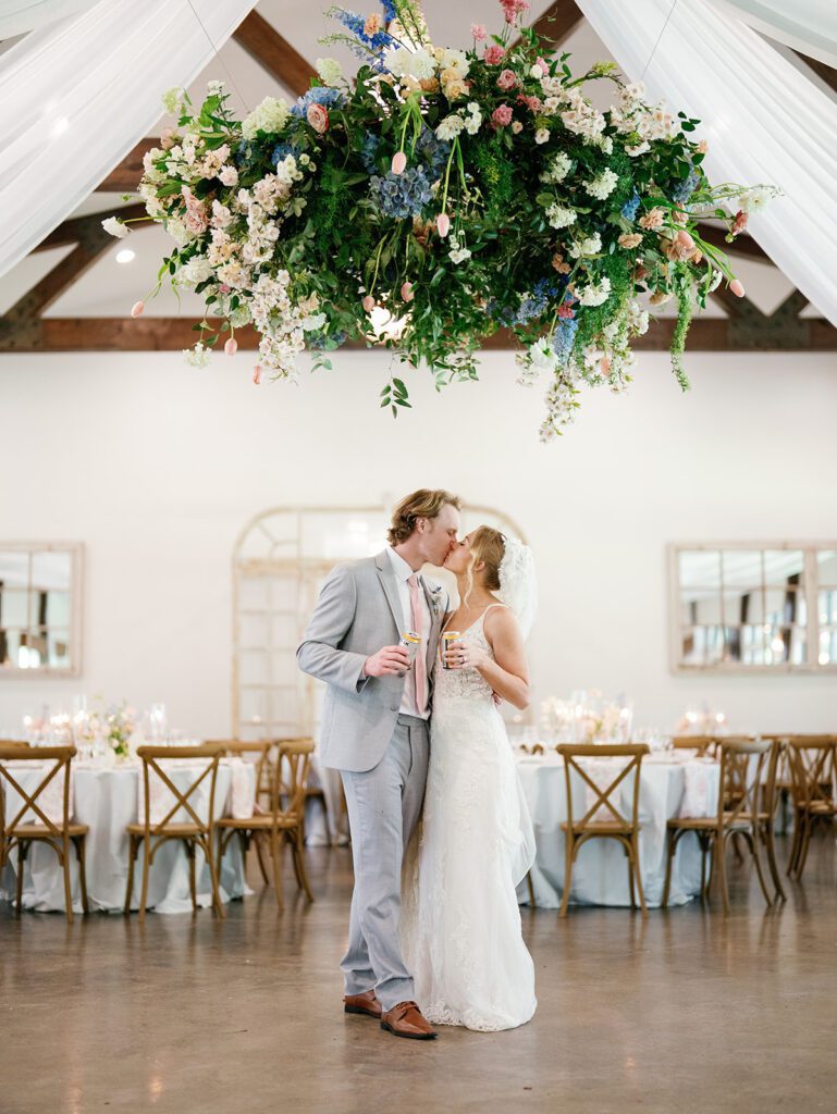 Bride and groom share a kiss beneath a floral chandelier wedding centerpiece, drinks in hand, with reception tables behind them