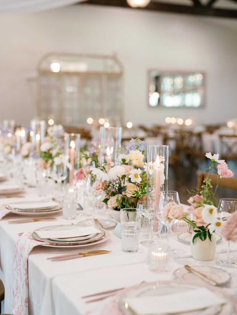 Long reception table with pastel bud vase arrangements, pink taper candles in glass hurricanes, gold flatware and pink floral napkins